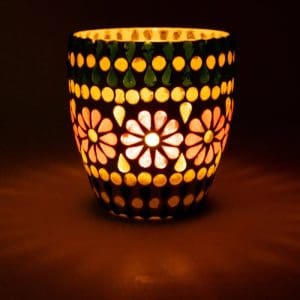 Цветен, осветен Мозаечен Стъклен Свещник зелено/жълто с флорални и точкови шарки хвърля топъл блясък в тъмна обстановка.