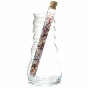 A wavy glass vase holds a corked test tube filled with small, colorful gemstones from the "От хаос до радост на живота" Пръчица за пречистване на вода, tilted at a slight angle.