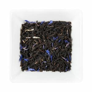 Бяла квадратна чиния съдържа насипни листа черен чай, смесени с малки сини венчелистчета от метличина, предизвикващи есенцията на Синя сянка.